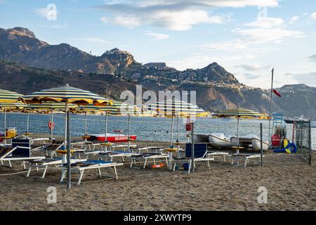 Leerer Strand mit gestreiften Sonnenschirmen und Liegen in Giardini Naxos, Sizilien. Atemberaubende Aussicht auf die Küste von Taormina und die Berge im Hintergrund. Beliebtes Ita Stockfoto