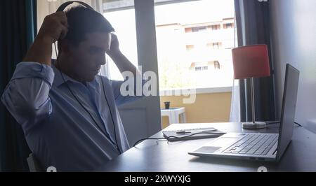 Ein junger Geschäftsmann zieht in seinem Heimbüro Kopfhörer auf, trägt ein blaues Hemd, um sich der Arbeit am Morgen zu stellen. Stockfoto