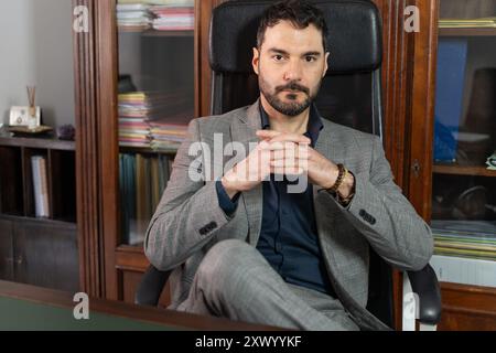 Reifer Geschäftsmann, der im Büro in Ledersesseln sitzt. Professioneller Mann mit Bart in grauem Anzug und blauem Hemd, der nachdenklich und selbstbewusst aussieht. Stockfoto