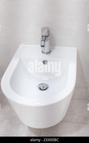 Bidet im Badezimmer. Gewöhnliches Badezimmer mit Waschbecken aus Keramik-Bidet. Weißes Bidet an eine beige geflieste Wand im Badezimmer geschraubt, Nahaufnahme, niemand Stockfoto