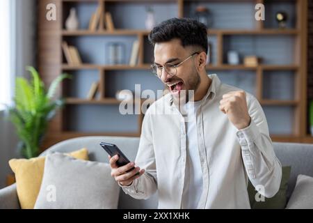 Junger indischer Mann, der zu Hause auf dem Sofa sitzt und auf den Bildschirm des Mobiltelefons blickt, glücklich mit Erfolg und erhielt Nachricht, die Hand zeigte ja. Stockfoto