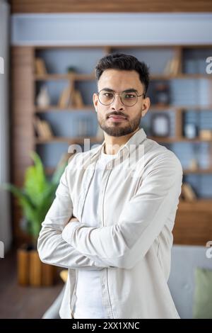 Porträt eines jungen Indianers mit Brille, der zu Hause steht, seine Arme kreuzt, ernsthaft steht und selbstbewusst in die Kamera schaut. Vertikales Foto. Stockfoto