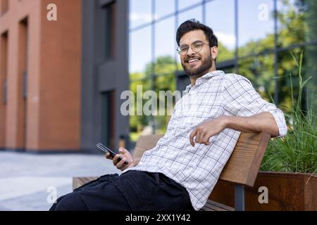 Ein junger Indianer sitzt entspannt draußen auf einer Bank und hält ein Telefon in der Hand. Schaut lächelnd in die Kamera. Stockfoto