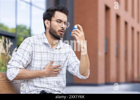 Junger Mann im Freien mit Asthma-Inhalator zur Atementlastung. Brille und kariertes Hemd tragen, Inhalator und Brust halten. Städtische Umgebung im Freien mit Grün und Gebäuden im Hintergrund. Stockfoto