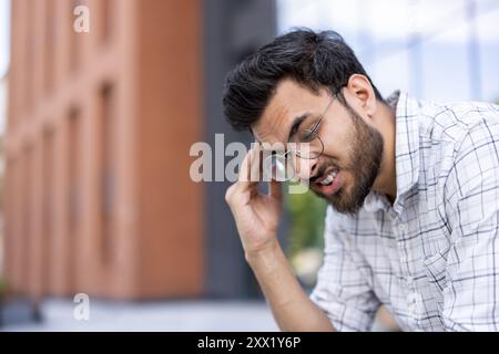 Junger Mann, der eine Brille trägt und im Freien Stress oder Müdigkeit erlebt. Der Mann hat eine Hand auf dem Kopf und zeigt Frustration oder Kopfschmerzen vor einem modernen Gebäude. Stockfoto