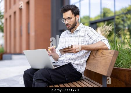 Junger Mann, der auf der Bank sitzt und Laptop und Telefon benutzt, während er Kopfhörer trägt. Reflektierendes modernes Bürogebäude im Hintergrund, legere Arbeitsplatzszene Stockfoto