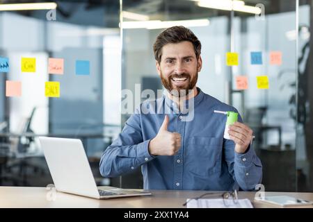 Porträt eines jungen lächelnden Mannes, der im Büro an einem Schreibtisch sitzt, ein medizinisches Spray mit Medikamenten in der Hand hält, auf ihn zeigt und in die Kamera lächelt. Stockfoto