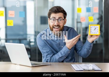 Porträt eines wütenden und emotionalen Geschäftsmannes, der im Büro am Schreibtisch sitzt, auf die Kamera blickt und auf die Uhr in der Hand zeigt. Stockfoto