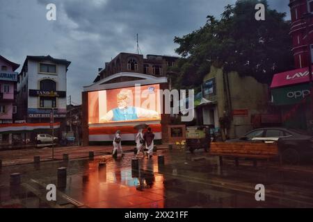 Srinagar, Indien. August 2024. Die Menschen laufen in einem Regen, während der indische Premierminister Narendra Modi am Straßenrand auf einem großen Videobildschirm zu sehen ist, der den Independent Day in Srinagar, dem indianischen Kaschmir, am Donnerstag, den 15. August 2024 feiert. (Foto: Mubashir Hassan/Pacific Press/SIPA USA) Credit: SIPA USA/Alamy Live News Stockfoto