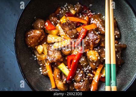 Schweinefleisch in süß-saurer Sauce in einer Schüssel auf dunklem Hintergrund mit chinesischen Essstäbchen und einer burgunderroten Serviette auf dunkelgrauem Hintergrund. Hochwertige Fotos Stockfoto