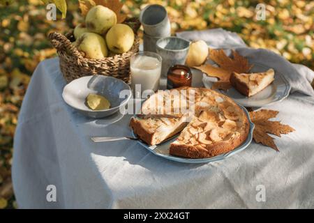Nahaufnahme eines Apfelkuchen und Tee mit einem Korb Äpfel auf einem herbstlichen Gartentisch in der Sonne Stockfoto