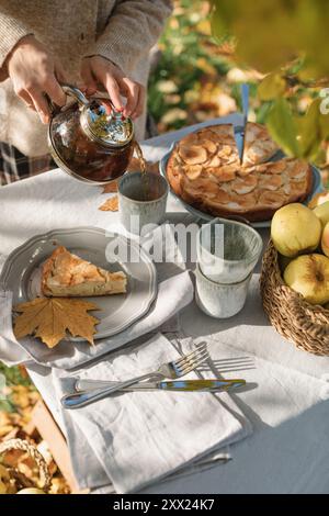 Eine junge Frau, die eine Tasse Tee und eine Tischunterlage mit Apfelkuchen und frischen Äpfeln auf einem herbstlichen Gartentisch in der Sonne gießt Stockfoto