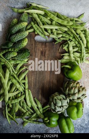 Aus nächster Nähe sehen Sie frische Artischocken, Stufenbohnen, Gurken und grüne Paprika rund um ein hölzernes Schneidebrett Stockfoto