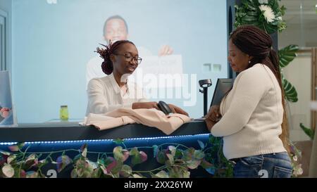 Frau mit Barcode-Scanner an der Kasse des Bekleidungsgeschäfts, die mit dem Kunden spricht, während sie darauf wartet, dass die gekauften Artikel verarbeitet werden. Retail Assistant scannt Kleidung, die der Kunde in einer Modeboutique gekauft hat Stockfoto