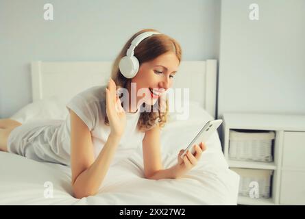 Glückliche, lächelnde junge Frau, die Musik mit kabellosen Kopfhörern hört und auf das Smartphone im weißen Zimmer zu Hause blickt Stockfoto