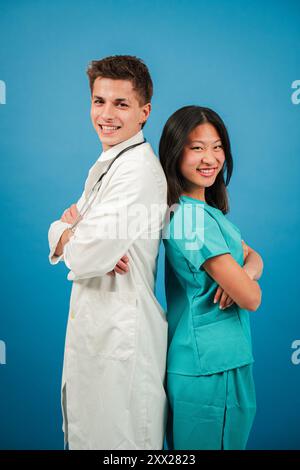 Zwei junge Erwachsene Ärzte, die auf Medizin spezialisiert sind, lächeln und blicken vor einer blauen Hintergrundwand in die Kamera. Das Team besteht aus einem Chirurgen, einem Kardiologen, einer Krankenschwester und einem Assistenzarzt. Hochwertige Fotos Stockfoto