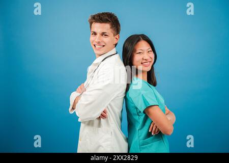 Zwei junge Erwachsene Ärzte, die auf Medizin spezialisiert sind, lächeln und blicken vor einer blauen Hintergrundwand in die Kamera. Das Team besteht aus einem Chirurgen, einem Kardiologen, einer Krankenschwester und einem Assistenzarzt. Hochwertige Fotos Stockfoto