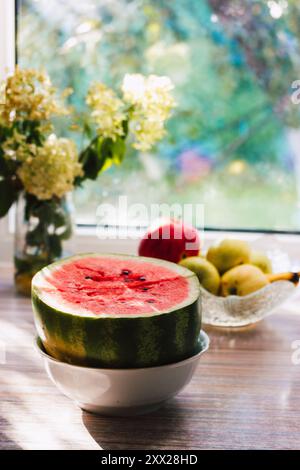 Stillleben mit Reifen Früchten auf einem Tisch in einer Landküche im Sonnenlicht. Wassermelone, Äpfel, Vase mit grünen Hortensien gegen Fenster in einem Landhaus Stockfoto