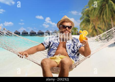 Ein Typ genießt in einer Hängematte mit einem Cocktail an einem exotischen Strand mit Wasservillen im Hintergrund Stockfoto