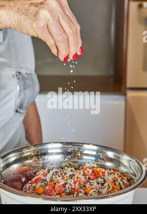 Nahaufnahme einer Person, die gemahlene Gewürze über einen bunten Salat in einer Edelstahlschüssel in der Küche streut. Stockfoto