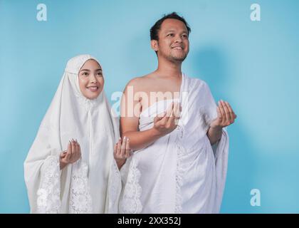 Lächelndes junges asiatisches muslimisches Paar, das ihram und Gebetskleid mit betender Handgeste trägt, die von blauem Hintergrund isoliert ist. Hajj-Konzept. Stockfoto