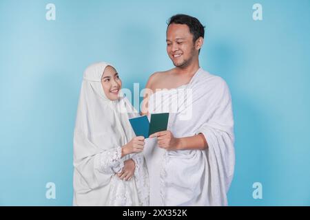 Junges asiatisches muslimisches Paar im Ihram und Gebetskleid, das einen Pass hält, während er sich ansieht, isoliert durch blauen Hintergrund. Hajj-Konzept. Stockfoto