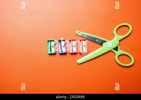 Eine Schere liegt auf einem Tisch mit vier verschiedenen Buntstiften. Die Bleistifte sind in einer Reihe angeordnet, wobei die grüne Schere sie durchschneidet. Stockfoto