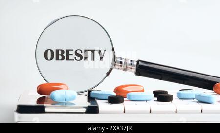 FETTLEIBIGKEITSWORT, geschrieben durch eine Lupe auf einem Rechner mit Tablets auf grauem Hintergrund. Der Begriff der Medizin Stockfoto