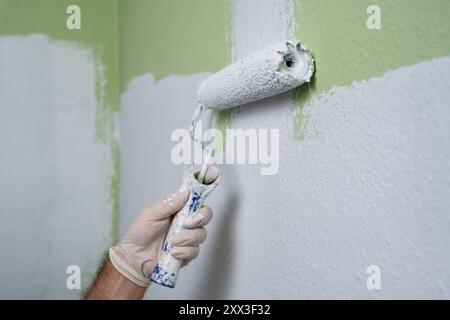 Die Hand eines Mannes malt eine grüne Wand im Zimmer mit einer Walze in weißer Farbe. Bauarbeiten und kosmetische Reparaturen im Haus, Wandmalerei, Tönung, Fini Stockfoto