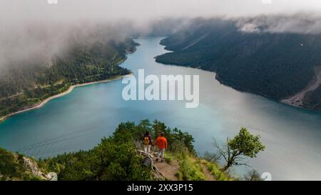 Zwei Wanderer genießen den atemberaubenden Blick auf den Plansee, der von üppigen Wäldern und sanften Bergen umgeben ist, während der Nebel langsam über Ihnen gleitet. Plansee Österreich Stockfoto
