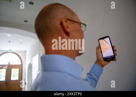 22. August 2024, Sachsen-Anhalt, Stendal: Falk Sichert, Geschäftsführer der Ingenieursgesellschaft Stendal, steht im Stendaler Rathaus und zeigt ein Programm auf einem Handy, das mittels Augmented Reality die Wand einer Domus Mercatorum, einer Markthalle aus dem frühen 12. Jahrhundert, an der Stelle zeigt, an der sie einst stand. Zwischen 2015 und 2016 wurden der Marktplatz und die angrenzenden Straßen im Rahmen eines Sanierungsprojekts archäologisch untersucht. Dabei wurden die Überreste des etwa 60 Meter langen Domus Mercatorum freigelegt. Das Gebäude kann nun virtuell mit Hilfe von erlebbar gemacht werden Stockfoto