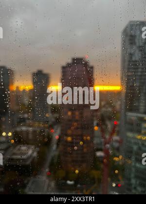 Dieses Foto zeigt einen verregneten Abend in Vancouver mit Regentropfen an einem Fenster, die den Blick auf die Skyline der Stadt bei Sonnenuntergang verwischen Stockfoto