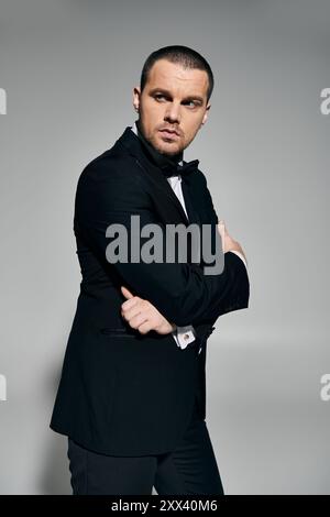 Ein Mann in einem schwarzen Smoking steht vor einem grauen Hintergrund, die Arme überkreuzt und zur Seite blickt. Stockfoto