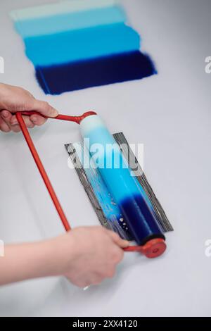 Hoher Winkel der Handwerkerhände, die am roten Griff der Walze mit blauer Farbverlaufsfarbe gehalten werden, während das neue Muster am Arbeitsplatz gedruckt wird Stockfoto