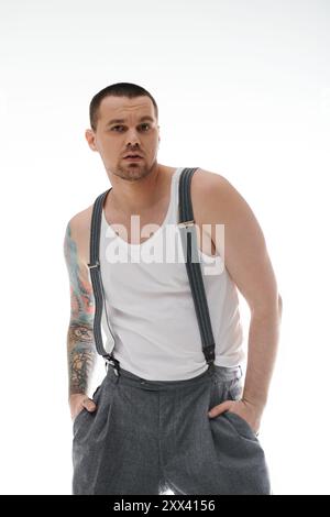 Ein Mann mit Tätowierungen am Arm posiert in einem weißen Tanktop und Hosenträgern. Stockfoto