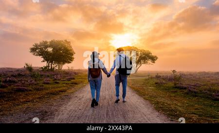 Ein Paar Mann und Frau geht Hand in Hand auf einem unbefestigten Pfad, umgeben von lebendigen Heidefeldern, während die Sonne hinter ihnen untergeht und ein warmes Leuchten über die Landschaft der Veluwe Niederlande wirft Stockfoto