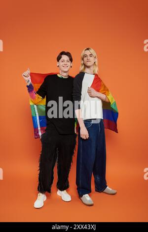 Zwei junge queere Freunde stehen zusammen, lächeln und halten eine Regenbogenfahne vor einem leuchtenden orangefarbenen Hintergrund. Stockfoto