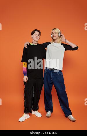 Zwei junge queere Freunde posieren in stilvoller Kleidung vor einem leuchtenden orangefarbenen Hintergrund. Stockfoto
