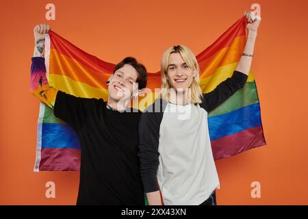 Zwei junge Freunde stehen lächelnd vor einem orangefarbenen Hintergrund und halten eine Regenbogenfahne hoch. Stockfoto