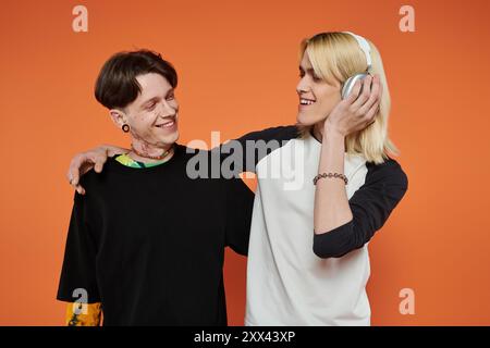 Zwei junge queere Freunde, in lebendiger Kleidung gekleidet, posieren zusammen vor einem leuchtend orangefarbenen Hintergrund. Stockfoto