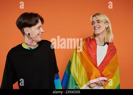 Zwei junge queere Leute mit stolzer Regenbogenfahne vor orangem Hintergrund. Stockfoto