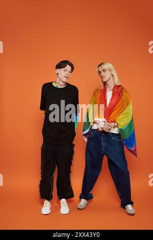 Zwei junge Freunde, die mit Regenbogenfahne stehen, posieren vor einem orangefarbenen Hintergrund. Stockfoto