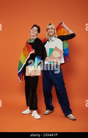 Zwei junge queere Freunde stehen stolz mit Regenbogenfahnen um ihre Schultern und halten Bücher vor einem leuchtend orangen Hintergrund. Stockfoto