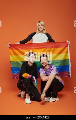 Drei junge Freunde, zwei kniend und einer stehend, mit einer Regenbogenfahne auf orangem Hintergrund. Stockfoto