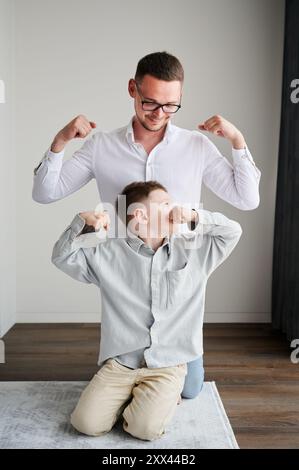 Der junge Vater spielt mit seinem neugierigen Sohn. Lustiger Junge, der seinem Vater Muskeln zeigt. Familie genießt Aktivitäten zu Hause. Stockfoto