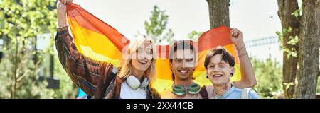 Drei junge queere Freunde stehen draußen zusammen, lächeln und halten eine Regenbogenfahne. Stockfoto