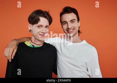 Zwei junge queere Freunde posieren zusammen in leuchtender Kleidung vor einem auffälligen orangen Hintergrund. Stockfoto