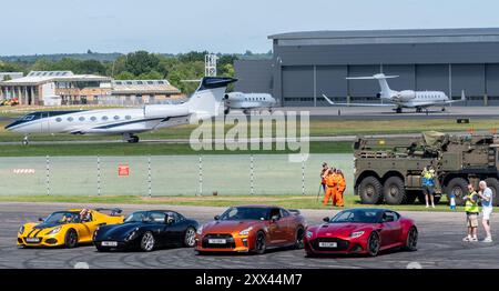 British Motor Show 2024, in Farnborough, Hampshire, England, Vereinigtes Königreich vom 15. bis 18. August 2024. Tag 2 der jährlichen Veranstaltung im Farnborough International Exhibition Centre. Blick auf Autos in der Arena mit Flugzeugen am Flughafen Farnborough im Hintergrund und Moderator Iwan Thomas. Stockfoto