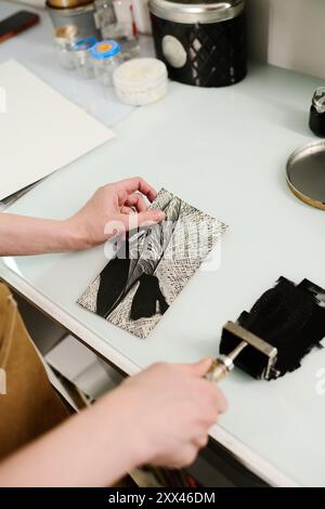 Hände junger, nicht erkennbarer Handwerker, die handgefertigte Kunstwerke und Pastorin halten, während sie schwarze Tinte über transparentes Glas verteilen Stockfoto