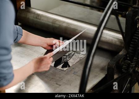 Hände einer jungen Designerin, die Papier auf ein Kunstmuster legt, während sie sich über eine Druckmaschine mit Metallschale und Walze beugt Stockfoto